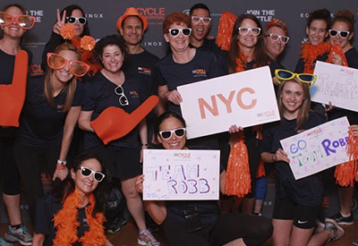 Members of TeamRobb holding signs, orange pom-poms, and orange foam fingers posing for a photo at a Cycle for Survival event.  