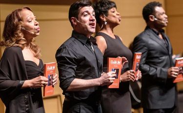 Four Visible Ink performers dressed in black hold orange program anthologies up to an audience as they sing.