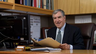 Man in suit smiling at desk, holding a folder, with bookshelves behind.